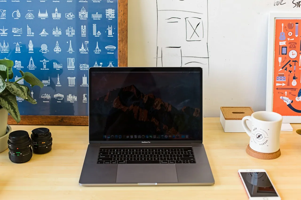 A MacBook Pro on a wooden desk beside a white mug, with a smartphone lying face-up at the front edge.
