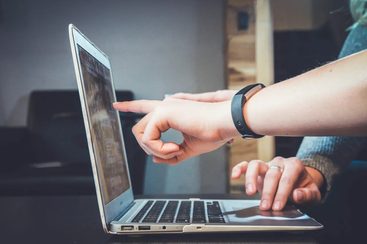 Two people's hands at a laptop, one pointing at the screen.