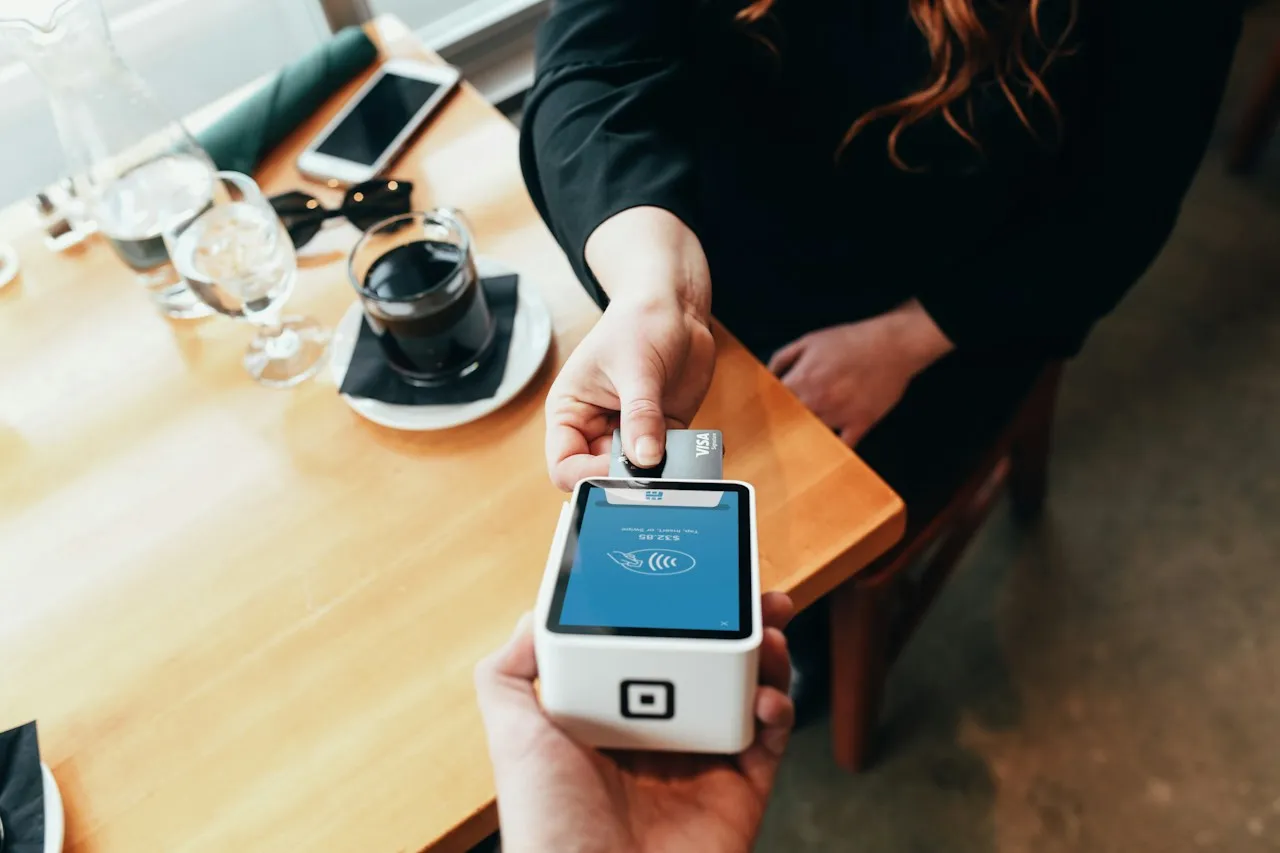 A customer inserts a card into a white card terminal held out by a staff member beside a café table with a glass of water, a coffee and a phone.