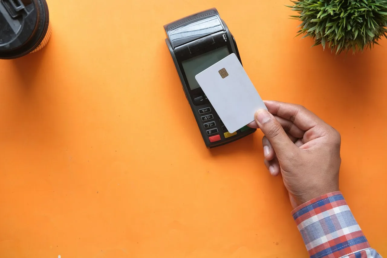 A hand holds a plain white card against a black card payment terminal on an orange tabletop.
