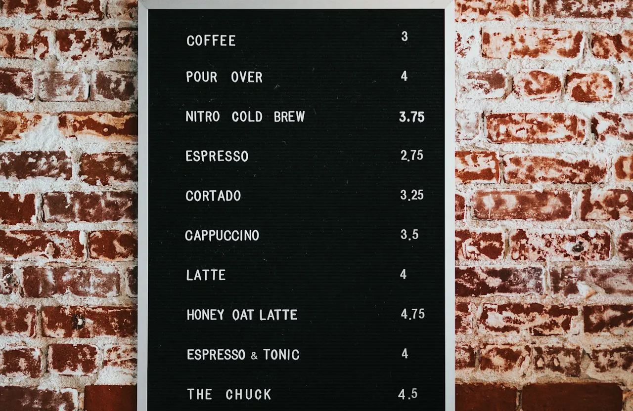 A black letter-board menu listing coffee prices on a whitewashed brick wall.