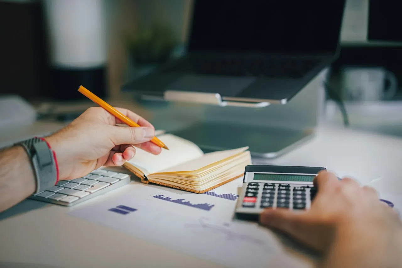 Hands at a desk, one holding a pencil over an open notebook and the other on a calculator, with printed papers and a laptop behind.