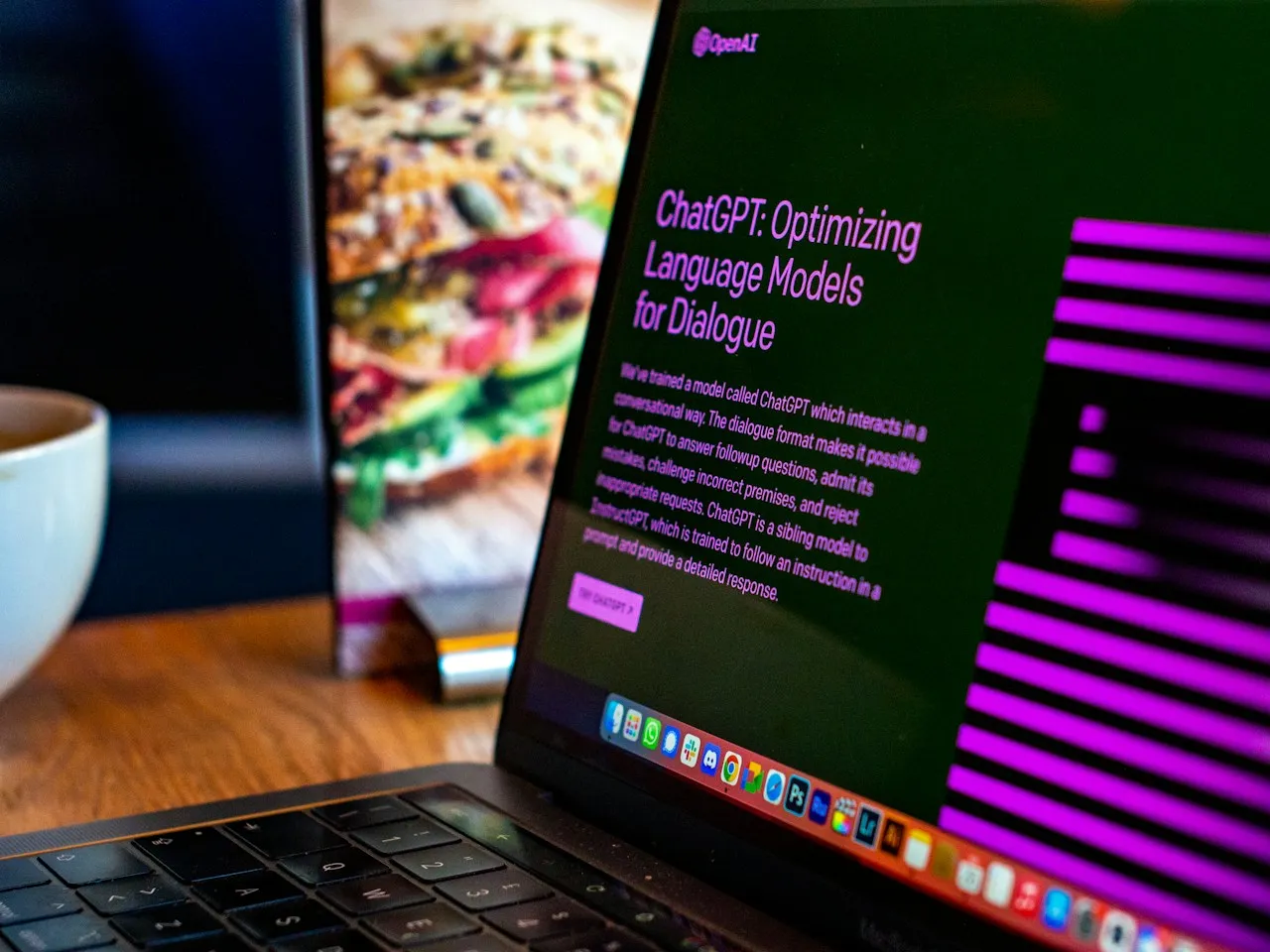 A laptop on a wooden desk shows OpenAI's ChatGPT web page, beside a coffee cup.