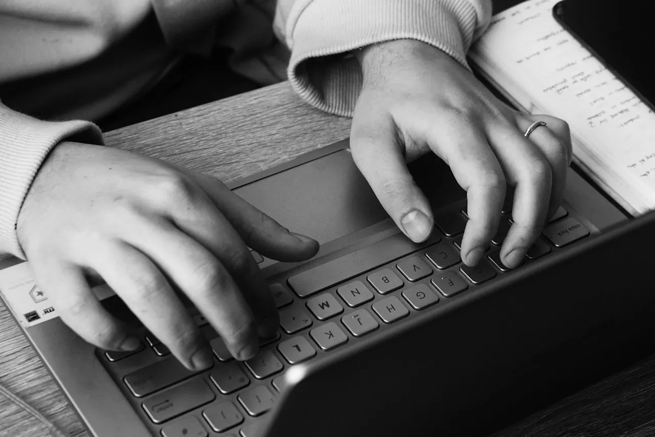 Black-and-white close-up of two hands typing on a laptop keyboard beside an open notebook.