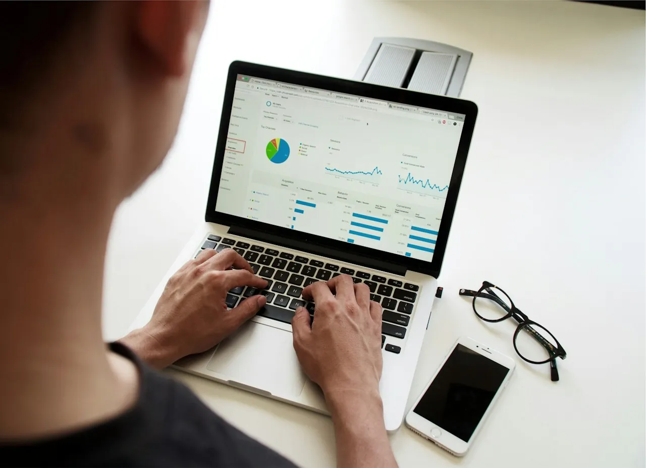 Seen over a person's shoulder, hands type on a laptop showing a marketing performance dashboard, with glasses and a phone on the desk.