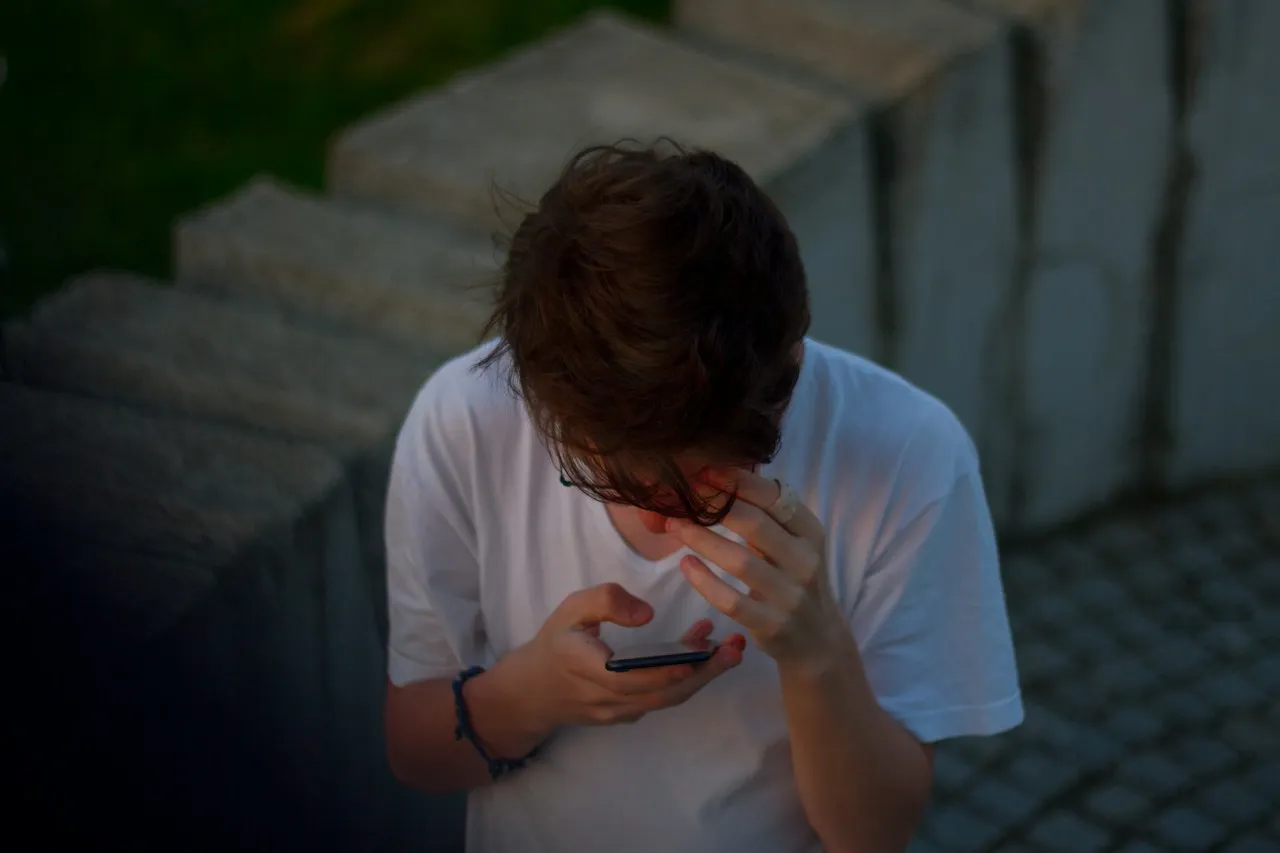A person in a white T-shirt looks down at a smartphone, one hand pressed to their forehead, their face hidden by their hair.