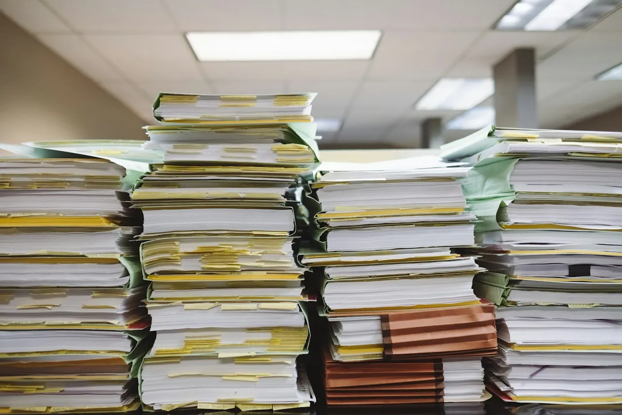 Tall stacks of paper documents and file folders on a desk in an office.