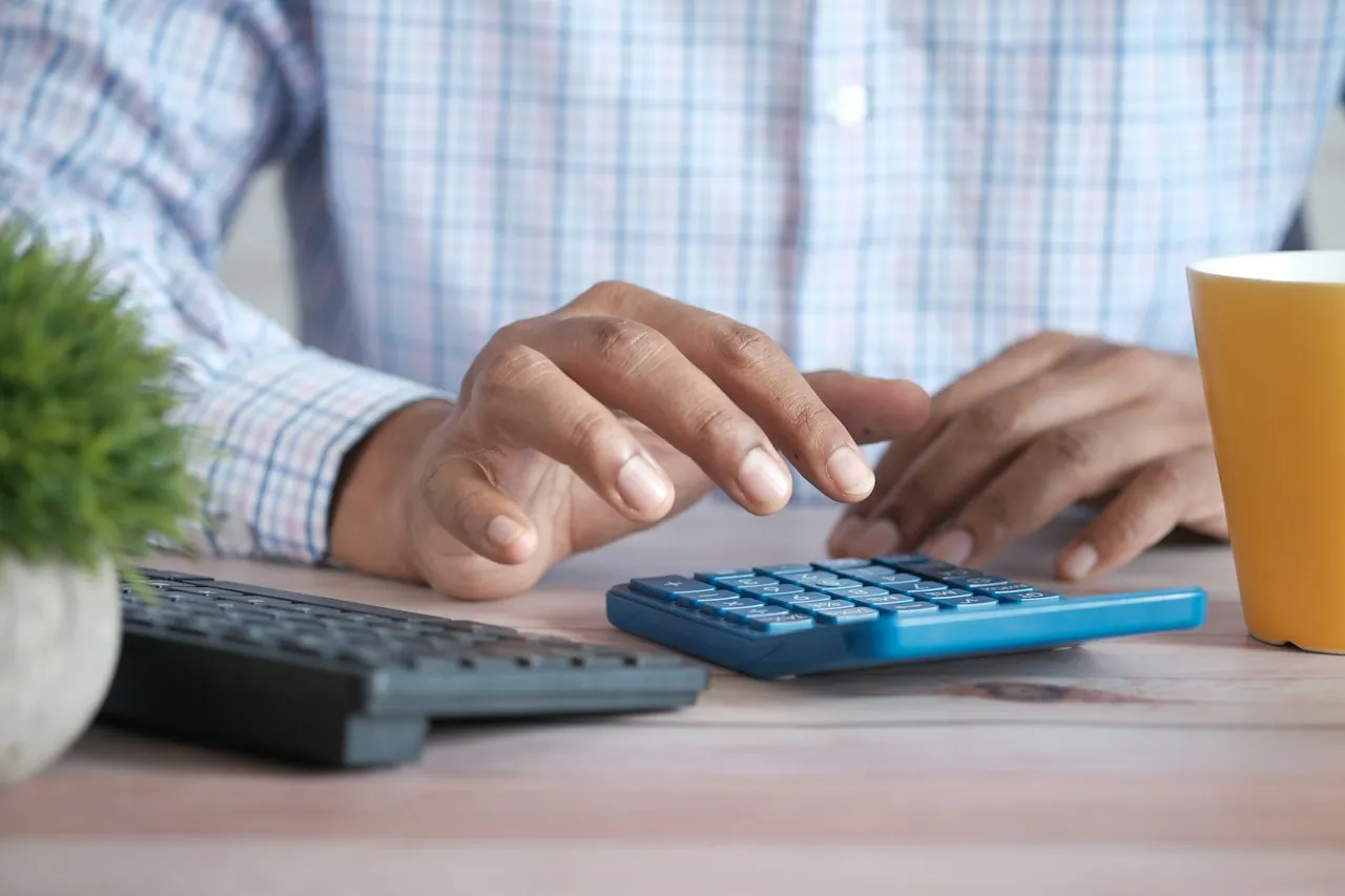 Hands work a blue calculator at a desk beside a keyboard, a yellow mug and a pot plant.