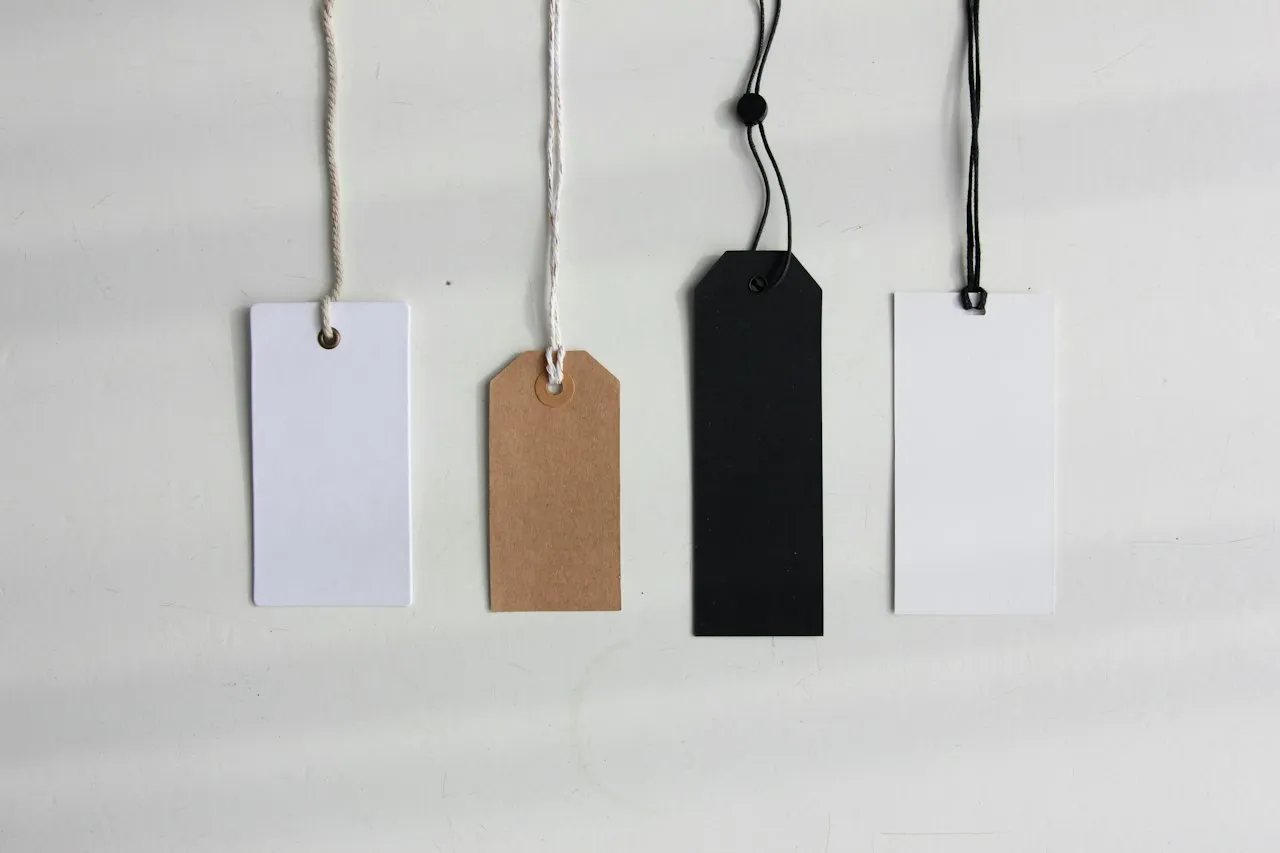 Four blank swing tags, white, brown kraft, black and white, hang on strings against a pale wall.