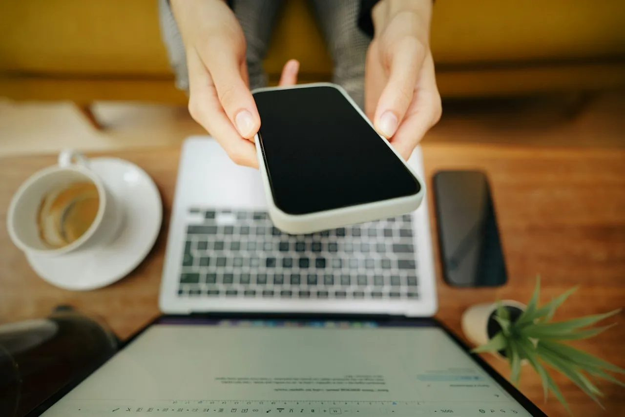 Seen from above, two hands hold a smartphone over a laptop keyboard on a wooden desk, beside a coffee cup, a second phone and a small pot plant.