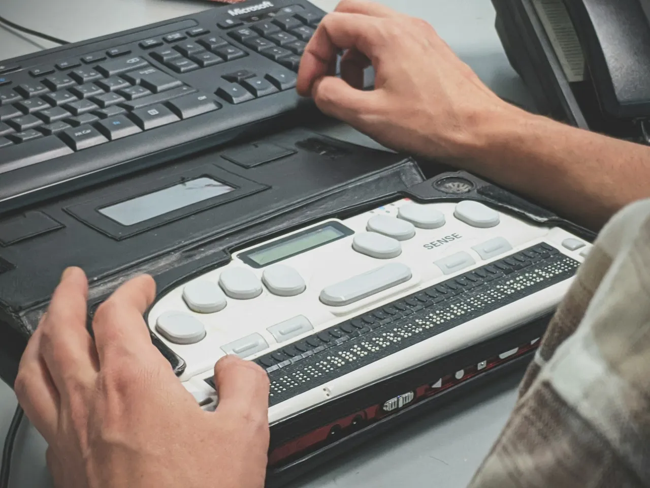 A person's hands rest on a refreshable braille display in front of a computer keyboard.
