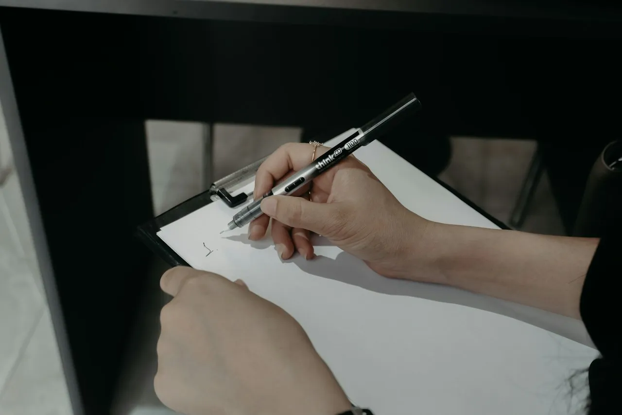 Hands writing with a pen on a blank sheet of paper held on a black clipboard.