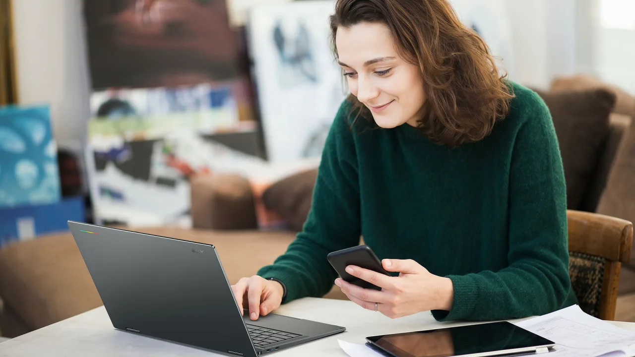 Acer's press image of a person at a table typing on an Acer Googlebook 14 with one hand and holding a smartphone in the other, with a tablet lying on the table beside the laptop.