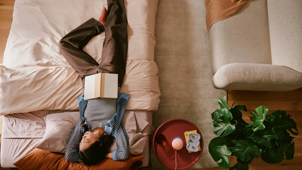 Amazon's press image, taken from above, of a woman lying on a bed reading a book, beside a small round red side table holding a pale pink Echo Dot.