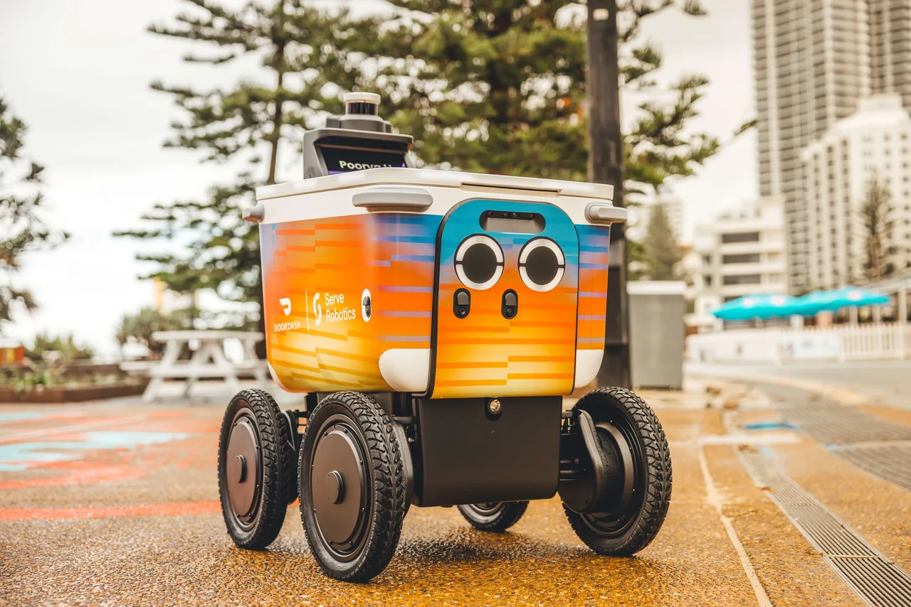 Press photo of a Serve delivery robot on a footpath, with Norfolk Island pines and high-rise apartments behind it. The robot is a box on four chunky black wheels, painted in an orange and blue gradient with DoorDash and Serve Robotics logos, two round camera 'eyes' on the front and a sensor on top.