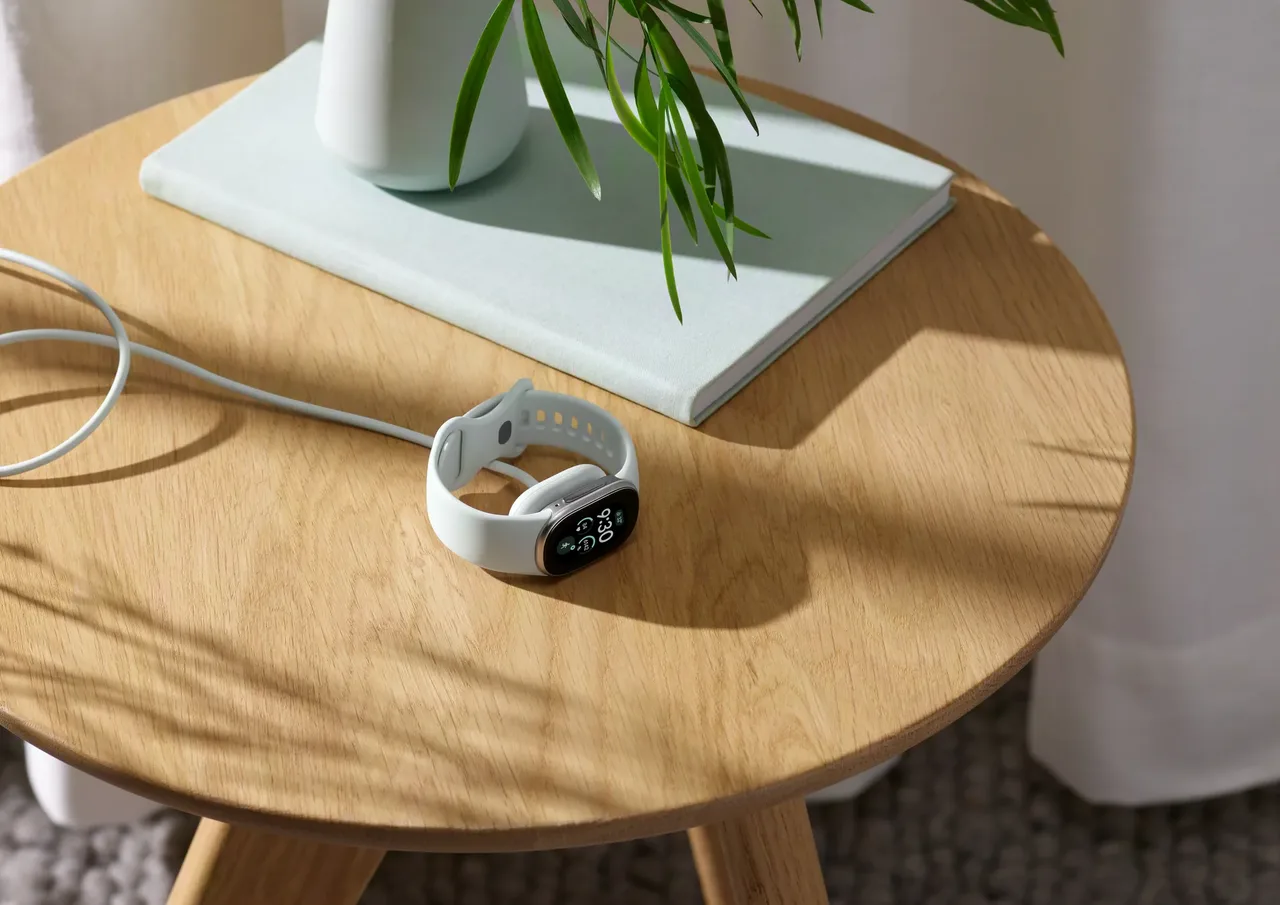 Google's press image of a Fitbit Edge with a pale band lying on a round wooden side table, connected to a white charging cable, its screen showing the time.