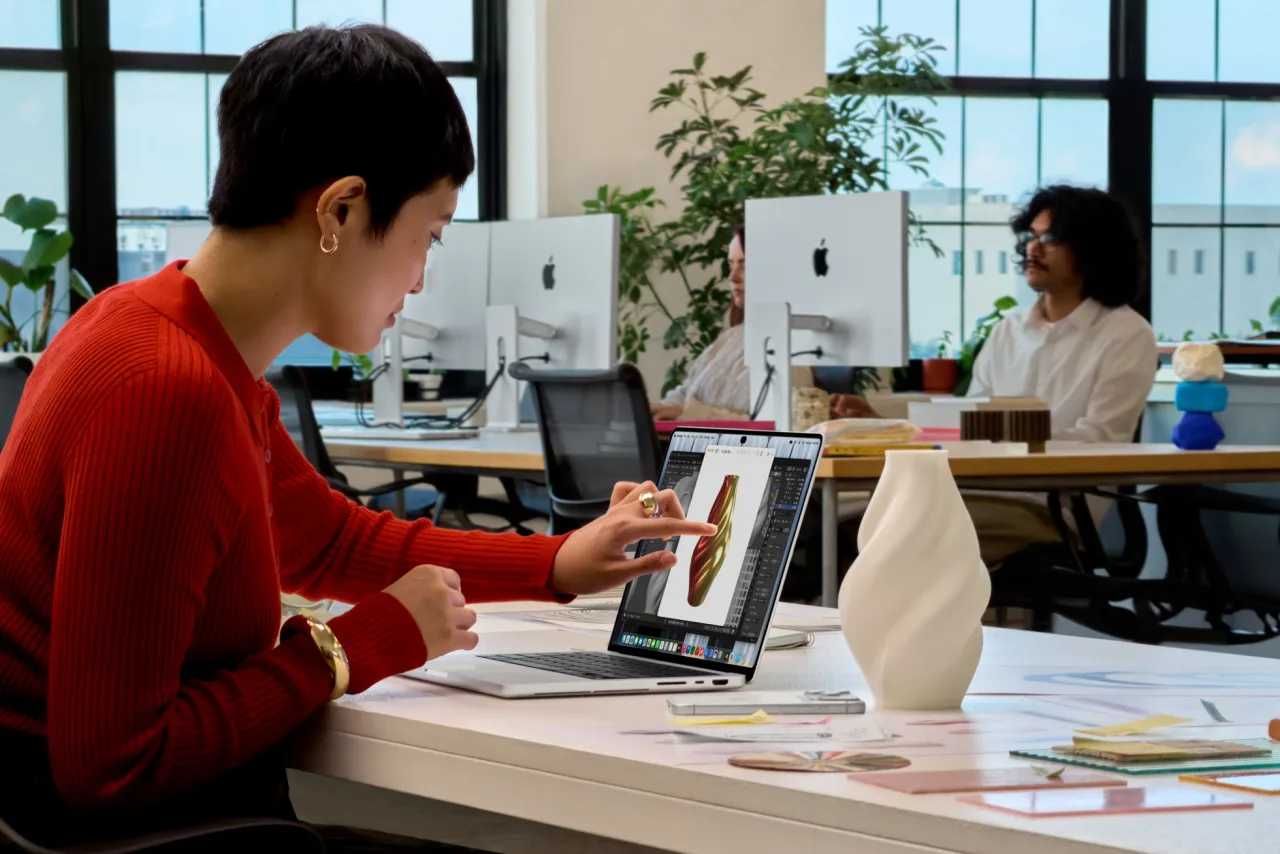 Apple's press image of a woman in a red jumper in a studio office, reaching out to touch a 3D model of a vase on the screen of a Silver MacBook Pro.